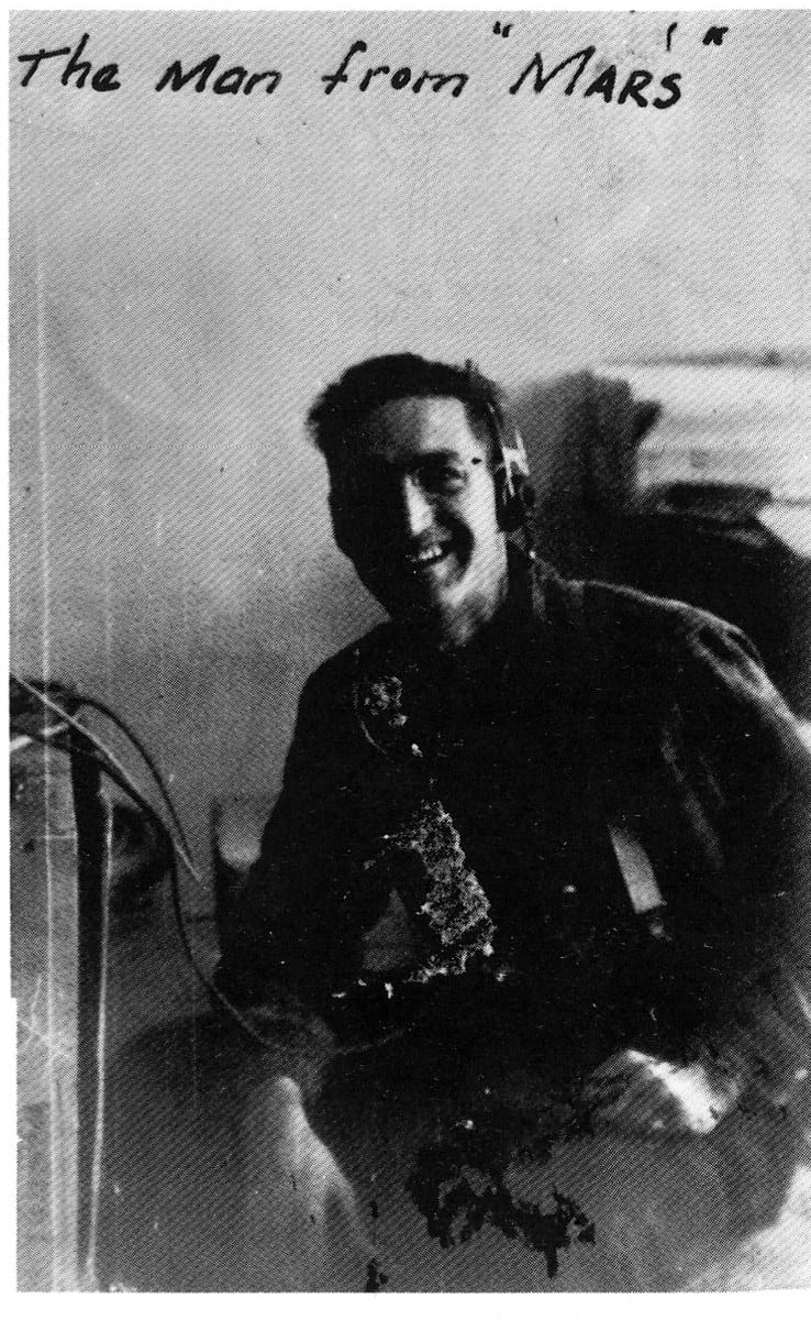 A grinning man in an operator's headset at a telephone switchboard, the photograph captioned by hand.