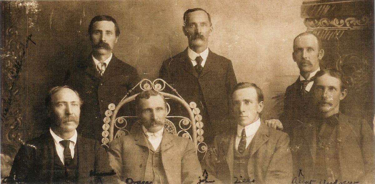Seven brothers in nineteenth-century suits posed for a studio portrait, three standing behind four seated.