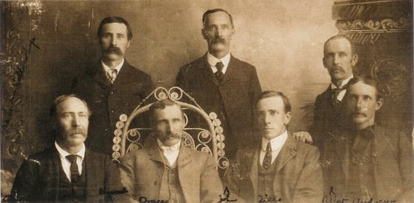 Seven brothers in nineteenth-century suits posed for a studio portrait, three standing behind four seated.